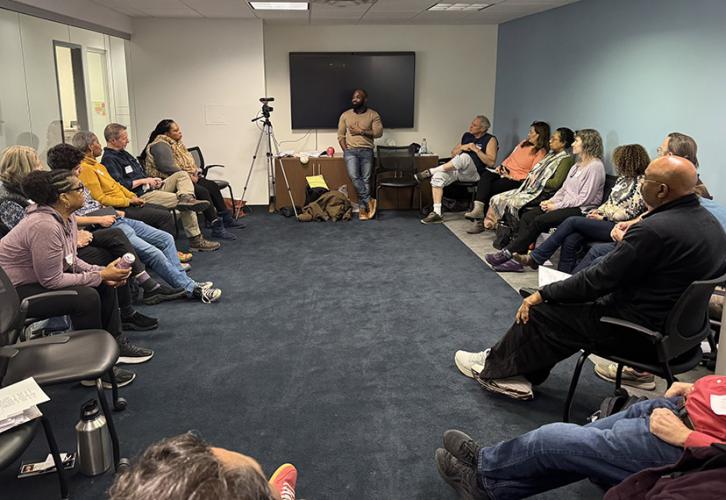 A large group of SAG-AFTRA New England members and staff sit in a circle of chairs in a conference room, angling themselves towards the TV screen to listen to the event’s guest speaker.