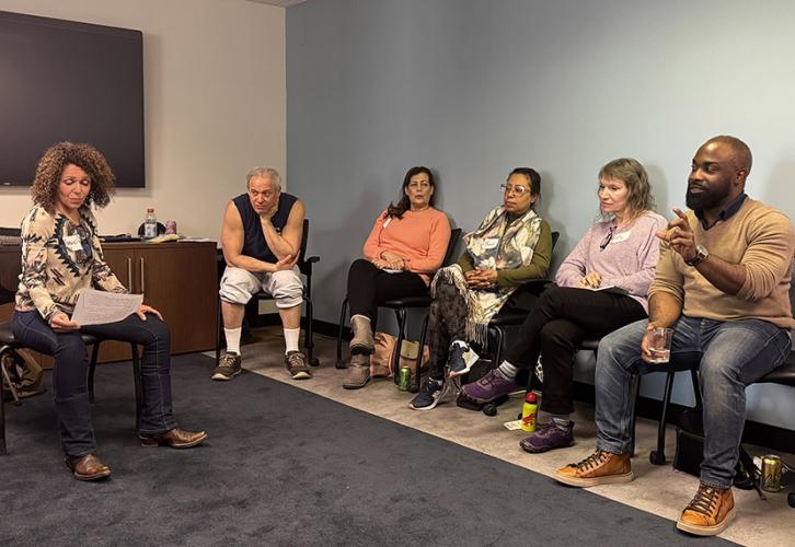 A group of five SAG-AFTRA New England members sit in chairs while participating in an acting exercise, one holding a piece of paper. The event’s guest speaker gestures while making a point.
