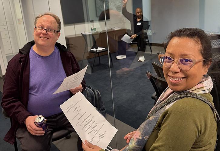 Pairs of SAG-AFTRA New England members sit in chairs; one pair in the foreground consists of a member in a red button-down and jeans and a member in a brown vest and black pants. Two more groups of two can be seen in the background further down the hallway.