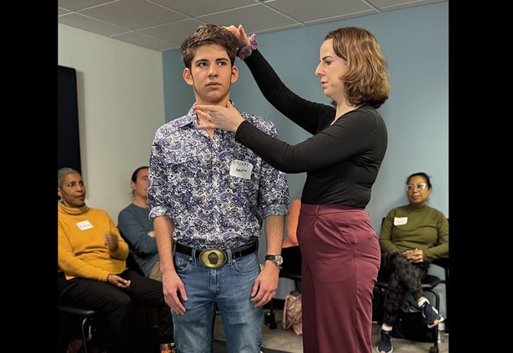An acting instructor in red pants and a black top adjusts a member’s posture by touching the top of his head and neck. Other members watch the demonstration in the background.