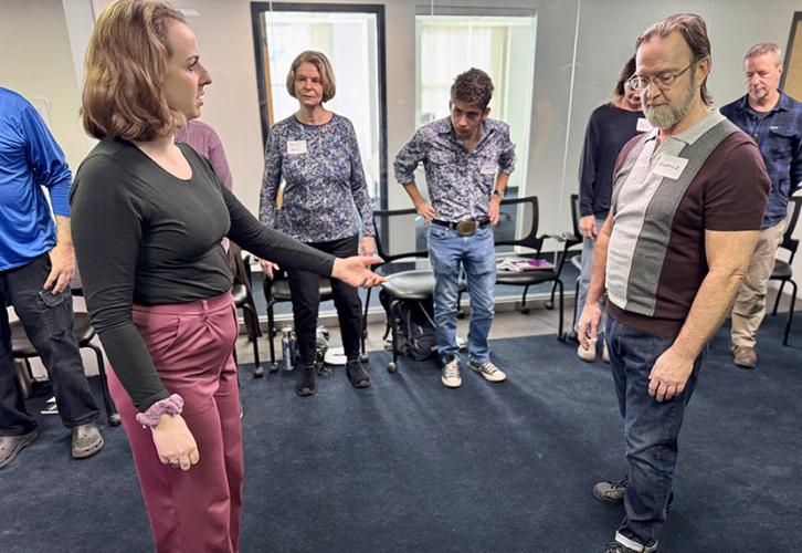 An acting instructor in red pants and a black top adjusts a member’s posture by touching the top of his head and neck. Other members watch the demonstration in the background.