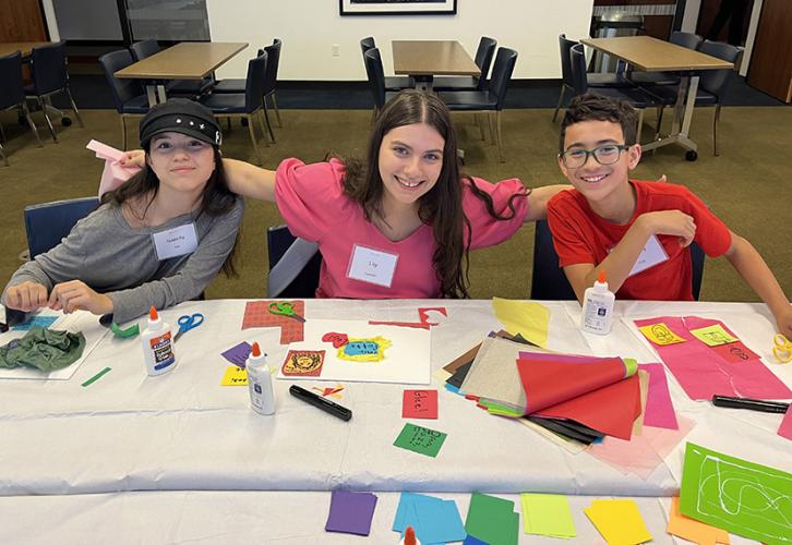 Three young performers sit at a table. On the table are art supplies, including glue bottles, scissors, canvases and brightly colored in different sizes. Behind them are brown tables and blue chairs and a piece of art on the wall with small headshots.