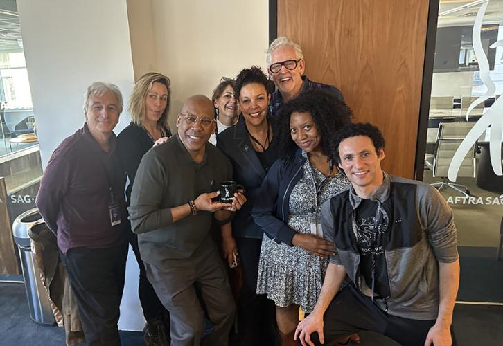  A diverse group of people smile for the camera in an office. The SAG-AFTRA logo is etched on a window next to them.