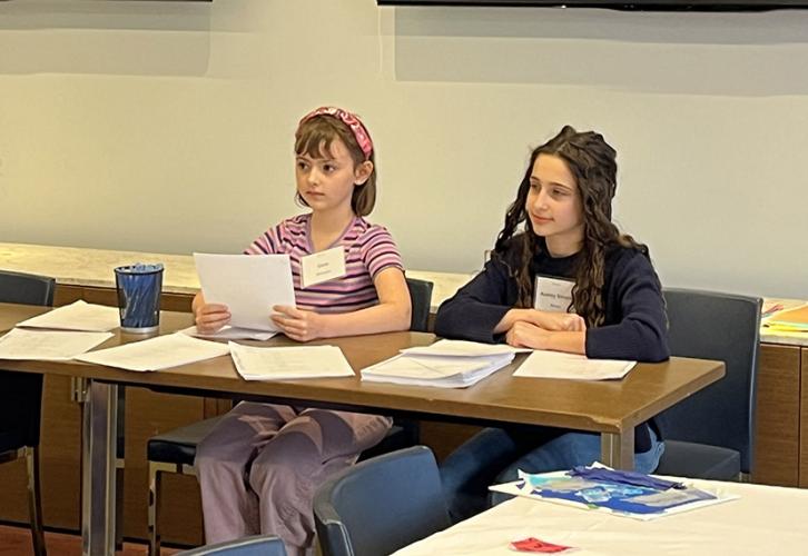 Two girls sit at a table. The one on the left holds a piece of paper. The one on the right sits with arms folded on the table. 