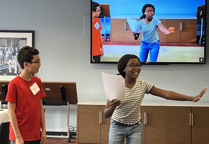 A boy in a red shirt stands looking at a girl on the right who is holding a piece of paper in one hand and holding out her arm. They  are also visible on the screen behind them.