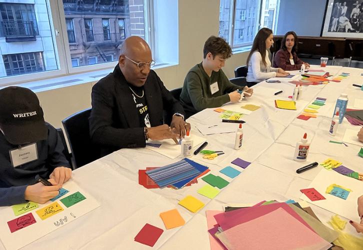 People sit at a table containing multicolored pieces of paper, glue, scissors, black markers, and canvases. They are creating collages with the materials available. 