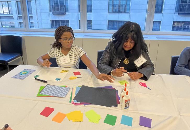 A girl and a woman sit at a table containing multicolored pieces of paper, glue, scissors, black markers and canvases. They are creating collages with the materials available. The girl, who is on the left, is reaching for supplies while the woman uncaps a black marker.