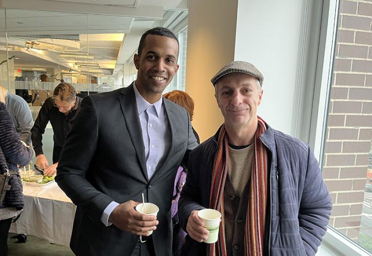 Two members stand next to a window holding coffee cups. Behind them are members in line getting food from the buffet. 