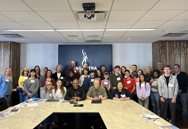 A group of children and adults stand behind a table in a conference room with the SAG-AFTRA logo on the wall. Five people are seated at the table.
