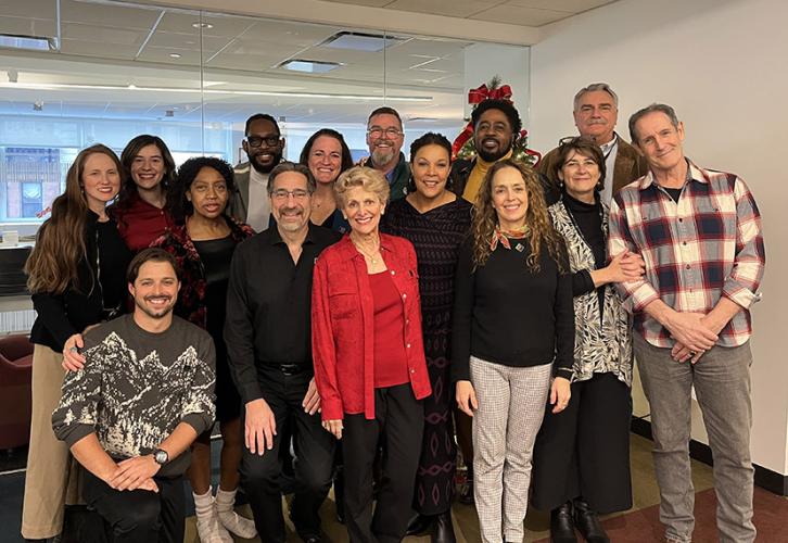 A diverse group of 15 people stand facing forward in an office. A Christmas tree is visible behind them.