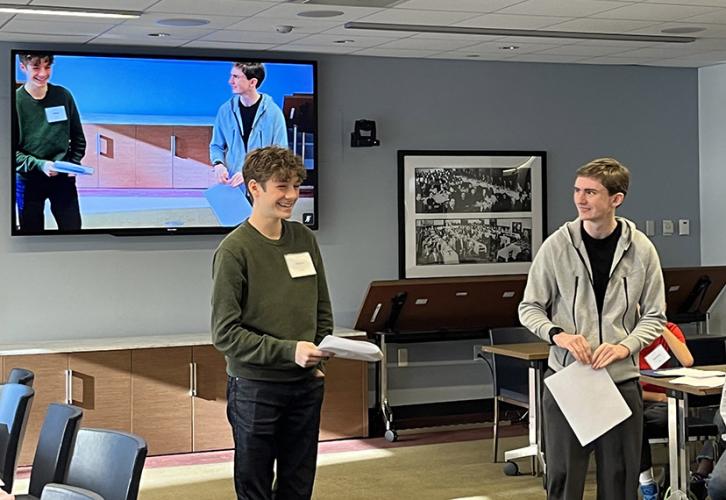 Two people stand in front of a screen where both of them are also visible. The person on the left is smiling and looking ahead. The person on the right is looking at the other person. One more person sits in a chair on the right side of the image. 