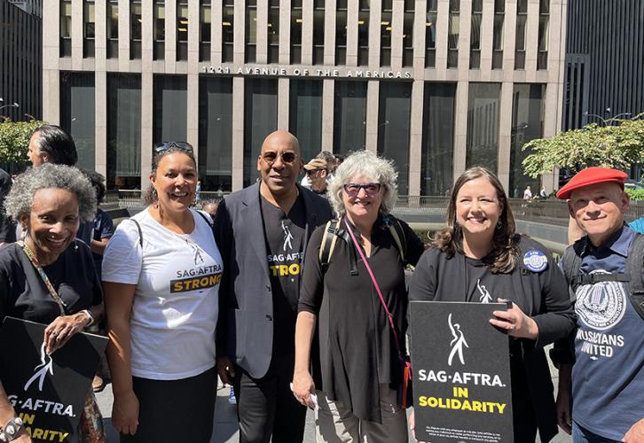 A group of six people stand facing the camera. One wears a white T-shirt with black and yellow text that reads SAG-AFTRA Strong, and one holds a sign with SAG-AFTRA In Solidarity written in white and yellow text on a black background.