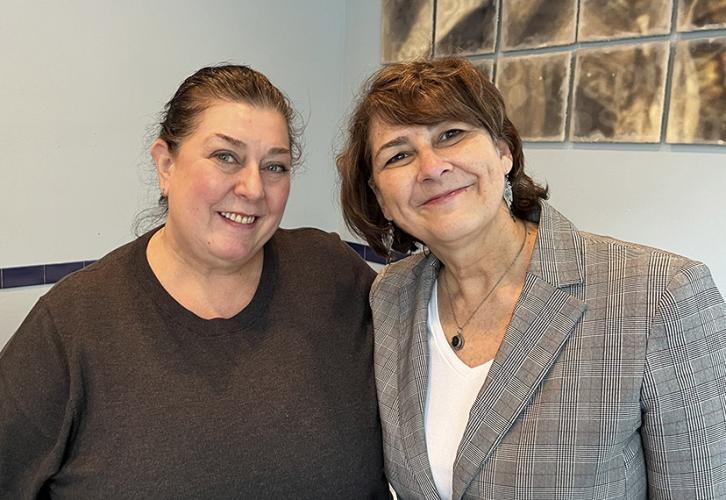 Two women smile for the camera. Behind them is a piece of beige and brown abstract art on a light blue wall. 