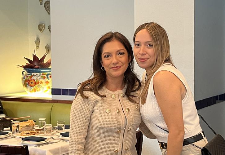 Two people pose for the camera in front of a light blue wall. Behind them to the left is a set table. 