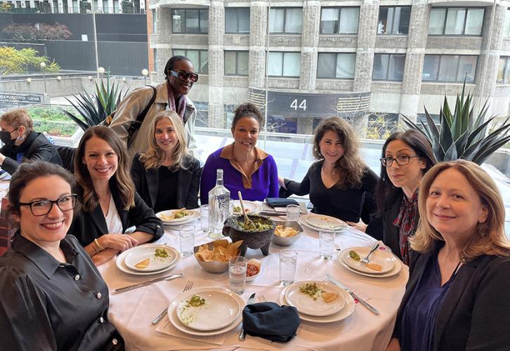 Seven women sit around a table at a restaurant while one stands behind them. A large building can be seen through the window behind them.