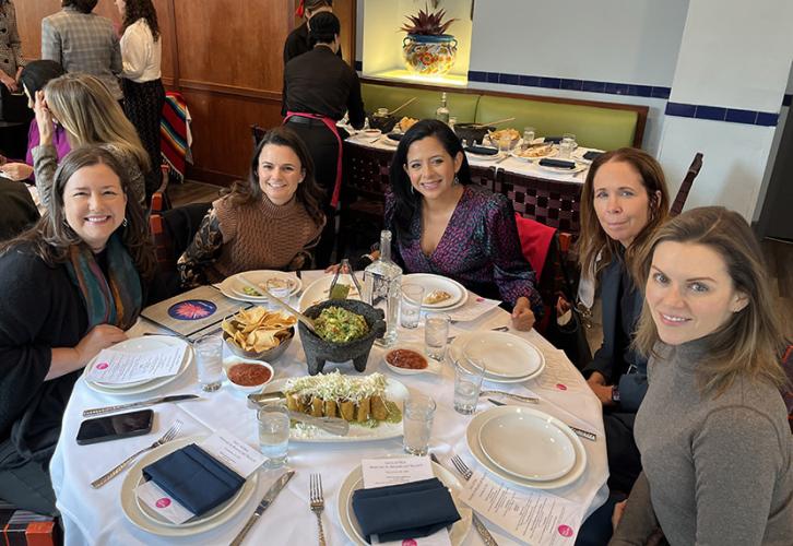 Five people sit at a round table. There is food at the center of the table. Behind them people stand in small groups speaking to each other.