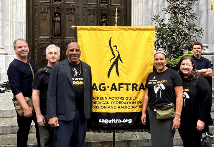 Five people stand in front of large carved brown cathedral doors on either side of a yellow flag with the SAG-AFTRA logo in black text. Beneath the logo reads Screen Actors Guild - American Federation of Television and Radio Artists. Below that in white text on a black background reads sagaftra.org.