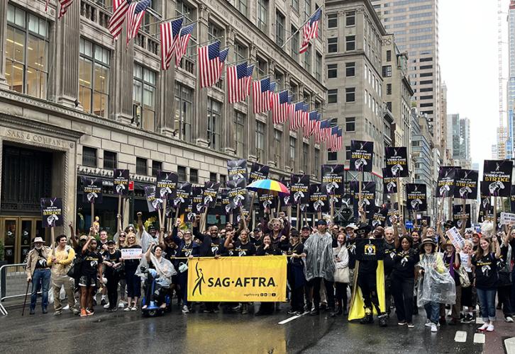 A large group of people stand on 5th Avenue in New York City. There is a row of American flags hanging from the building behind them. They hold SAG-AFTRA Video Game Strike picket signs with white and yellow text on a black and purple background. The people at the front of the group hold a SAG-AFTRA parade banner with black text on a yellow background.
