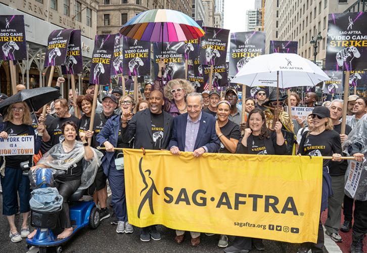 A large group of people stand behind the SAG-AFTRA parade banner with black text on a yellow background. One person holds a large umbrella with rainbow-toned panels. Another holds a white umbrella with a black SAG-AFTRA logo. Others hold SAG-AFTRA Video Game Strike picket signs with white and yellow text on a black and purple background.