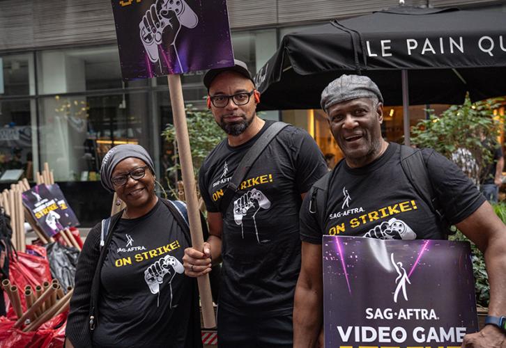 Three people are wearing black SAG-AFTRA On Strike T-shirts and hold SAG-AFTRA Video Game Strike picket signs with white and yellow text on a black and purple background.