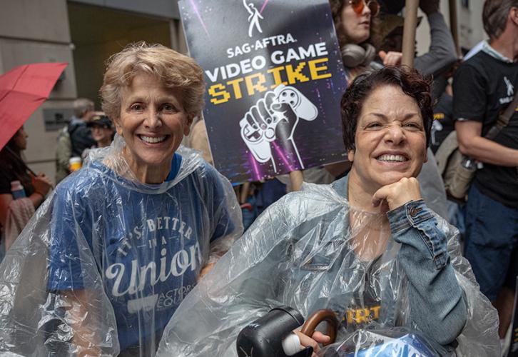 Two women stand in front of a SAG-AFTRA Video Game Strike picket sign with white and yellow text on a black and purple background.