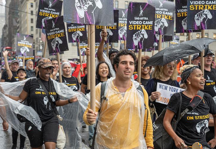 A group of people move along a street holding SAG-AFTRA Video Game Strike picket signs with white and yellow text on a black and purple background.