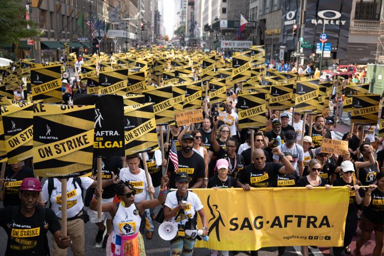 Un grupo grande y diverso de personas camina por una calle de la ciudad con carteles de piquete de SAG-AFTRA y una gran pancarta amarilla de SAG-AFTRA.