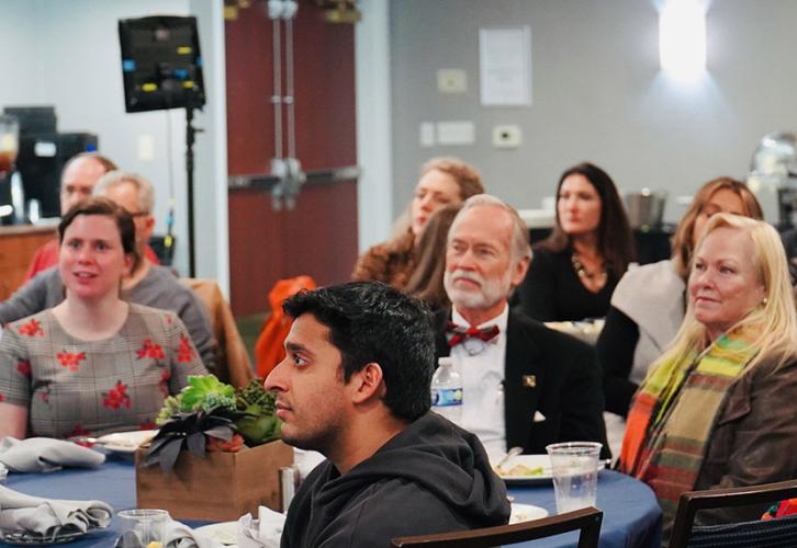 People sit around round tables in a multipurpose room, listening to a speaker who is not pictured.
