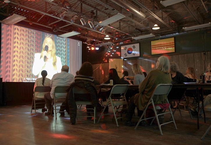 Seated people in a darkened room watch a large projection screen. On the screen a blond woman in a white suit addresses the audience.