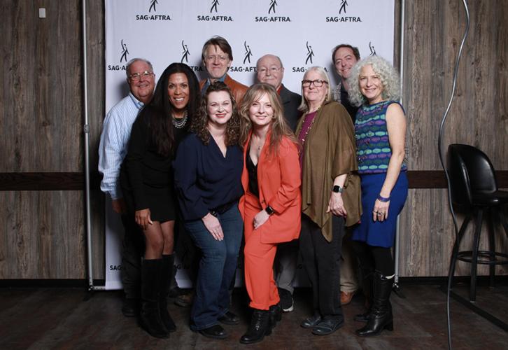 A group of nine well-dressed people smile for a photograph in front of a white SAG-AFTRA step-and-repeat banner.