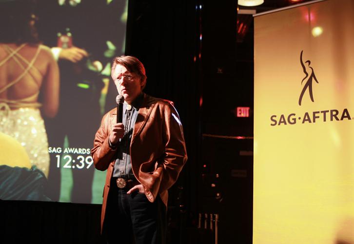 A man in a brown leather jacket holds a microphone. Behind him is a large projection screen and a yellow SAG-AFTRA banner.