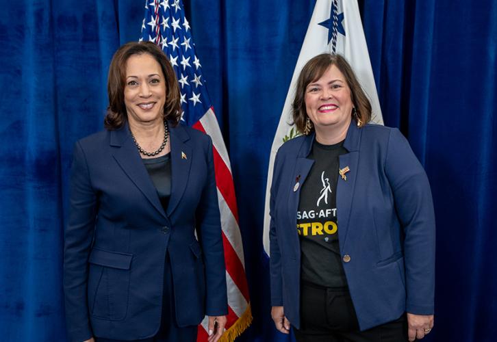Harris, left, wearing a blue suit, stands next to Maher, who is wearing a blue jacket over a SAG-AFTRA Strong T-shirt. Behind them is a blue backdrop and two flags, including an American flag. 