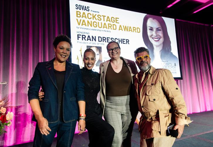 Powell, Baker, Johnson and Stivale stand closely together. Behind the group on a projector screen is a title card bearing Fran Drescher’s headshot. Other text includes “Backstage Vanguard Award” in gold lettering; and Fran Drescher in black lettering.