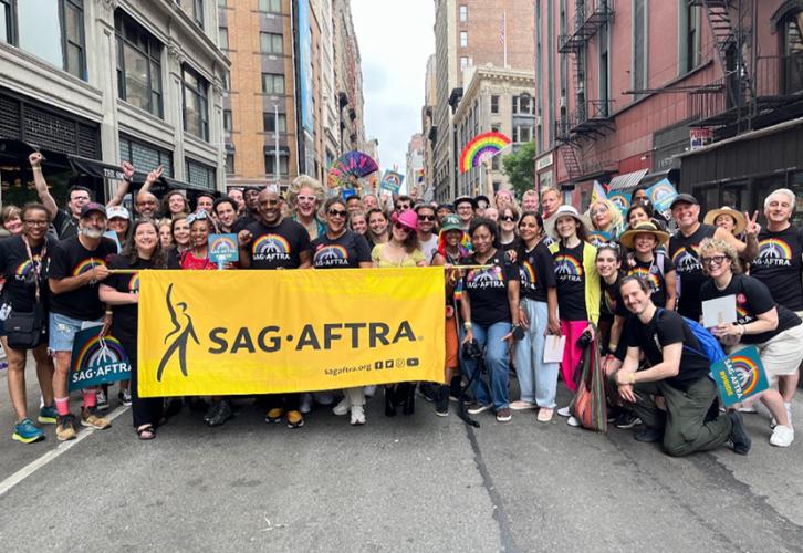 Un gran grupo de personas se reúne detrás de una pancarta amarilla con un logotipo negro de SAG-AFTRA en el centro. La gente usa camisetas negras con un arcoíris y el logo blanco de SAG-AFTRA en el centro. Algunos tienen abanicos de color verde azulado con un logotipo de SAG-AFTRA en blanco y arcoíris.