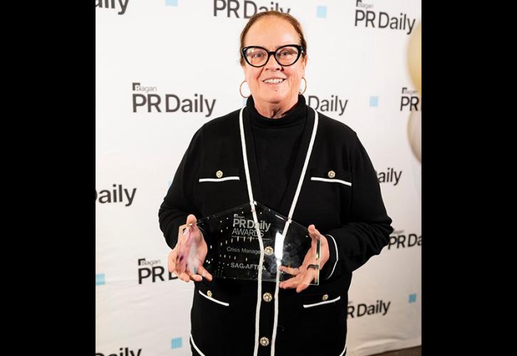 Greenwalt se encuentra de pie sobre la alfombra roja frente a un fondo blanco con el logo de PR Daily. Lleva un suéter negro con ribetes blancos y botones dorados y una camisa negra de cuello alto. En sus manos hay una estatuilla de vidrio con un texto en blanco.