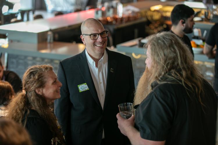 Escobedo, Taylor and Braa chat with one another. Escobedo and the man are seen in profile; Taylor faces the camera. Braa holds a glass in his hand. 
