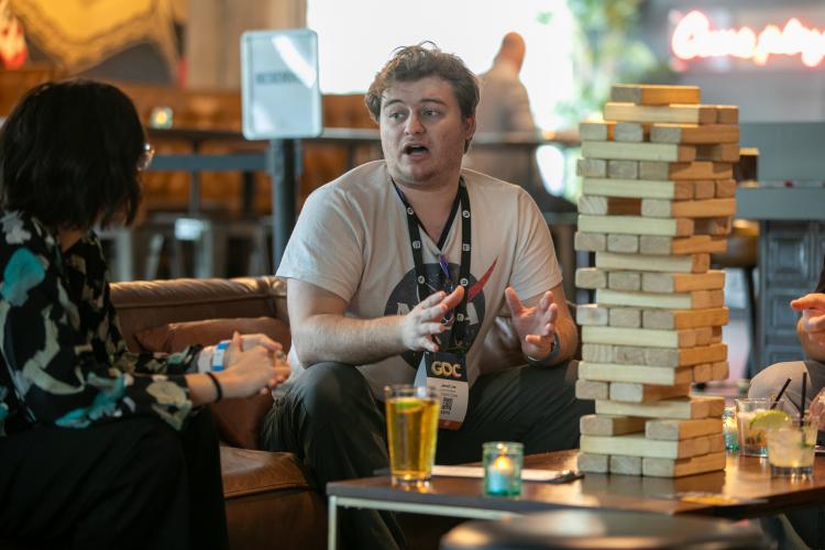 A man, center, is prominently displayed. He turns towards a woman with short black hair who faces away from the camera. In the foreground is a Jenga game with large blocks.