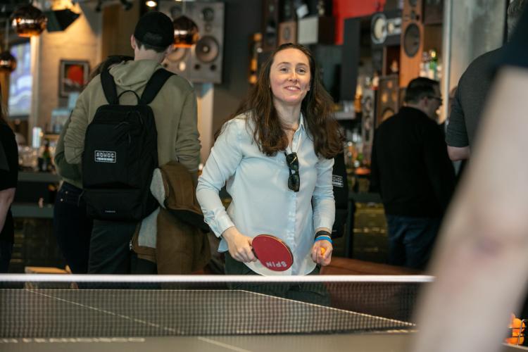 A woman in a denim blue shirt is prominently seen behind a ping pong table. She holds a red ping pong paddle in her right hand and a ping pong ball in her left. She looks towards her unseen opponent.