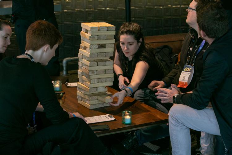 Woodward, center, is seated. She leans forward to take a Jenga block from a game. She wears a black t-shirt and bracelets on both arms. Others around her are Elmaleh and Crabtree-Ireland.
