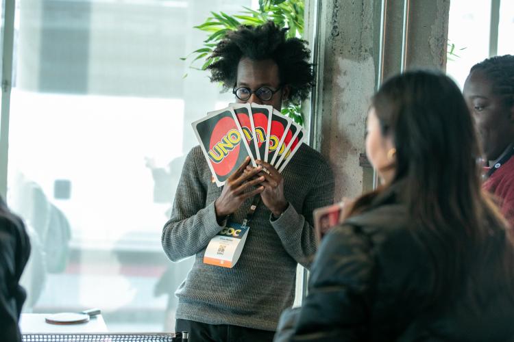 An African American man is prominently displayed holding large Uno cards. The back of the cards, black and red with “Uno” in yellow copy, are seen. To the lower right are two other people.