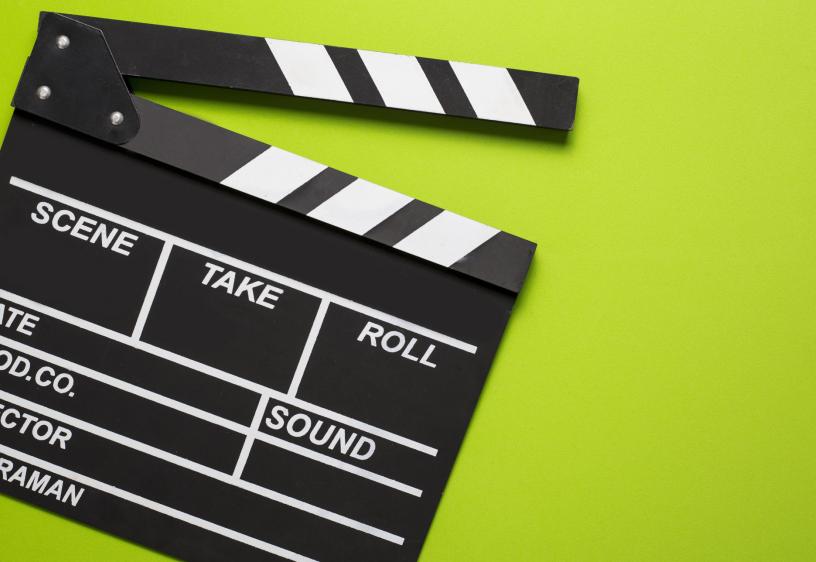 A black and white clapperboard is placed on a green background. The clapperboard is slightly open and has sections labeled "Scene", "Take", "Roll", "Date", "Prod Co", "Sound", "Director", and "Cameraman."