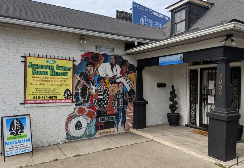 White and black building with a painted colorful mural depicting musicians playing instrumnets such as a guitar, saxophone, and drums. A sign on the wall provides information about the museum, including its hours and contact details. 