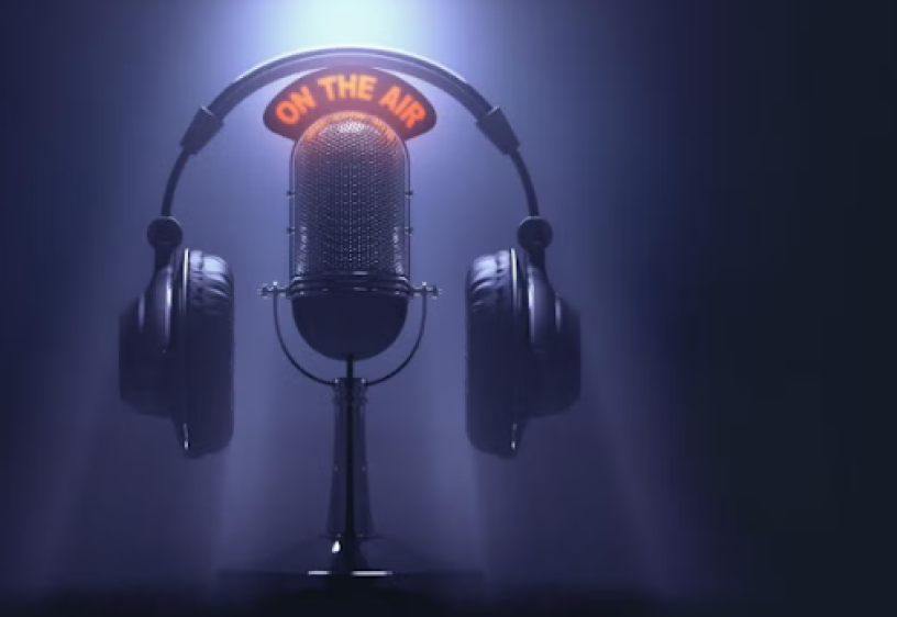 A black radio microphone stands with "on the air" in red wording above it. On top of the "on the air" sign is a pair of black headphones. The microphone, "on the air" sign and headphones appear in a spotlight in front of a black background.
