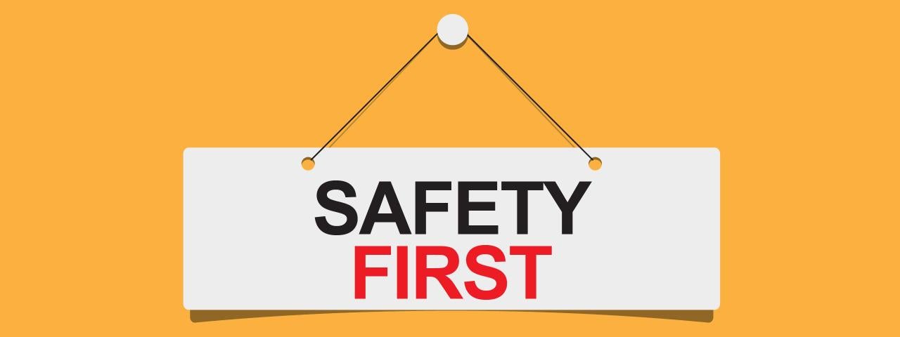 Opening Soon "SAFETY" in black "FIRST" in red cursive on a white panel hanging from a whitepin with black string on a dandelion yellow background