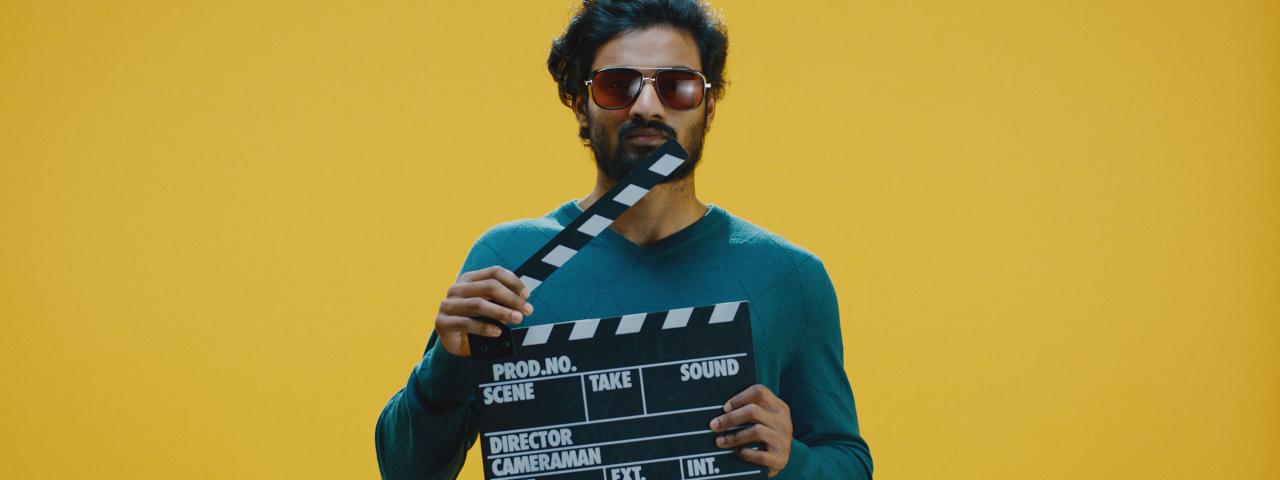 Image of a gentlemen holding a clapperboard wearing a green long sleeve and wearing dark shades standing in front of a yellow background.