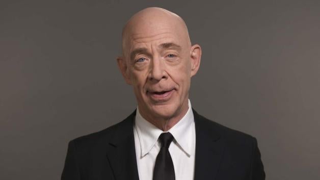 Simmons in a black suite with a white shirt and a black tie standing in front of a grey backdrop