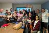 Un grupo diverso de 16 personas se paran y se arrodillan en dos filas para una foto grupal. Muchos sonríen mientras sostienen varios equipos de práctica de RCP, incluidos dos maniquíes de RCP para bebés y dos maniquíes de RCP para adultos, uno de los cuales está vestido con una camiseta negra de SAG-AFTRA. Horrigan, en la fila de atrás, sostiene un maniquí de RCP para adultos sobre su cabeza.