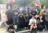 Un grupo diverso de 19 personas que visten una variedad de camisetas de SAG-AFTRA e IATSE se paran y se arrodillan con un arco de globos blancos, azules y naranjas en el fondo y una pancarta blanca alta a la izquierda que dice en letras azules y naranjas: "Es mejor en un sindicato". Una de las personas está de pie con su perro atado con una correa.