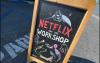 A photo of an outdoor blackboard-style sign in a parking lot. The hand-designed sign reads “Netflix and the Workshop” in all caps, with “Netflix” in red. There is also a cartoon sketch of a boy falling into bed as various characters watch his descent.