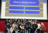 Una foto de un grupo grande y diverso en el escenario debajo de una pantalla de cine. Muchos visten camisetas solidarias negras de SAG-AFTRA. Sobre ellos, en la pantalla de cine, está la copia: "¿Qué es #AdsGoUnion?" y cuatro espacios en blanco debajo.